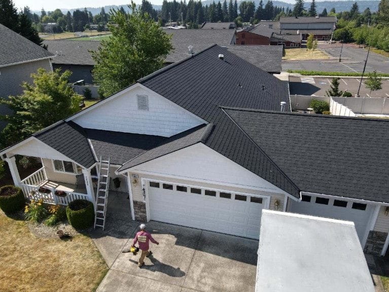 Aerial view of completed asphalt shingle roof replacement on single-story home with roofer on driveway